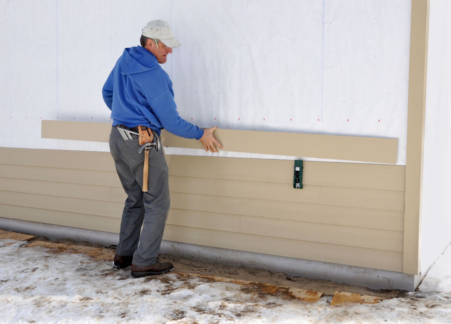 A person in a blue hoodie installing tan lap siding on the exterior wall of a building.