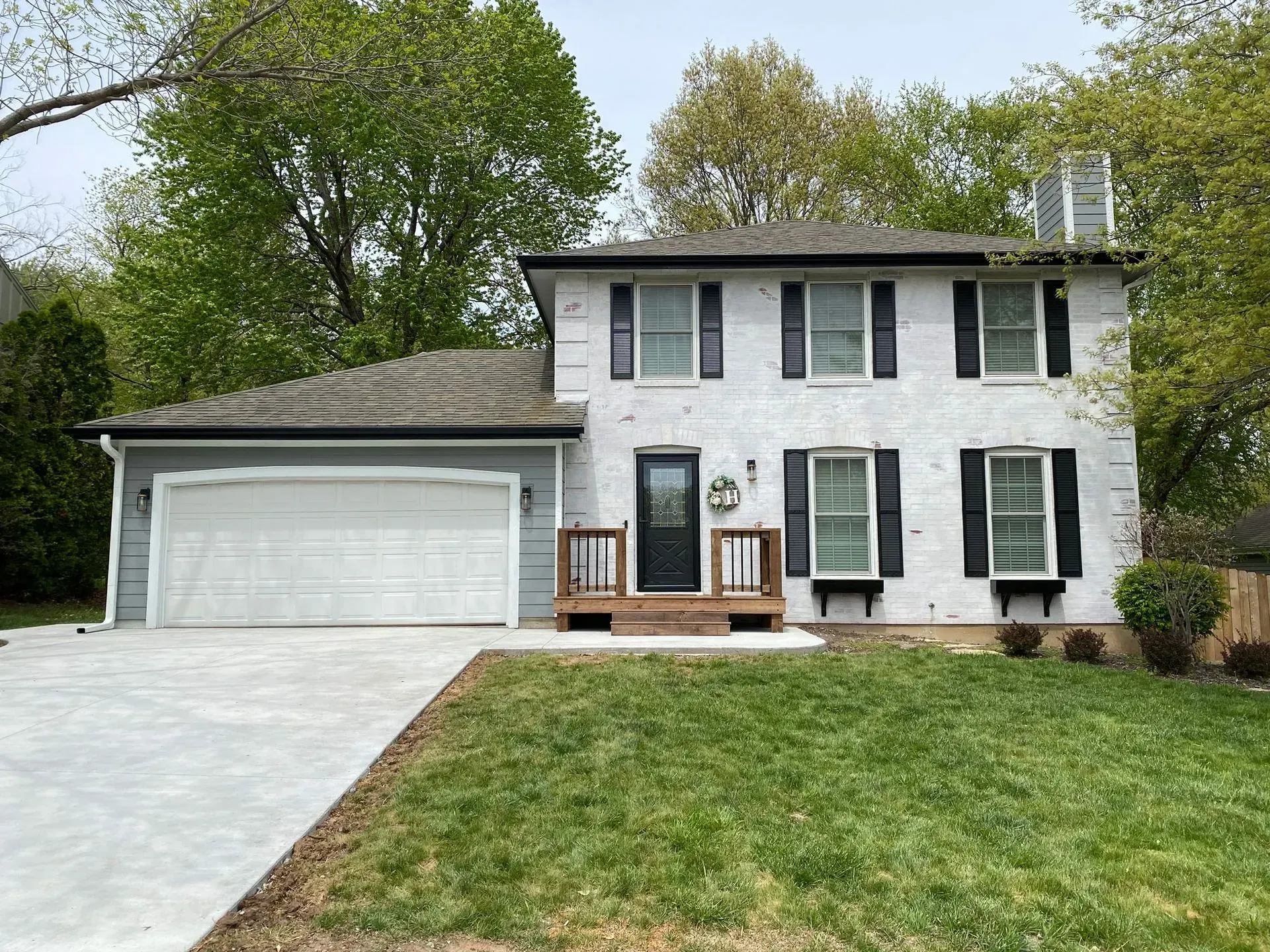 A two-story house with a white painted brick exterior, gray garage, black shutters, and a front porch, set among trees.
