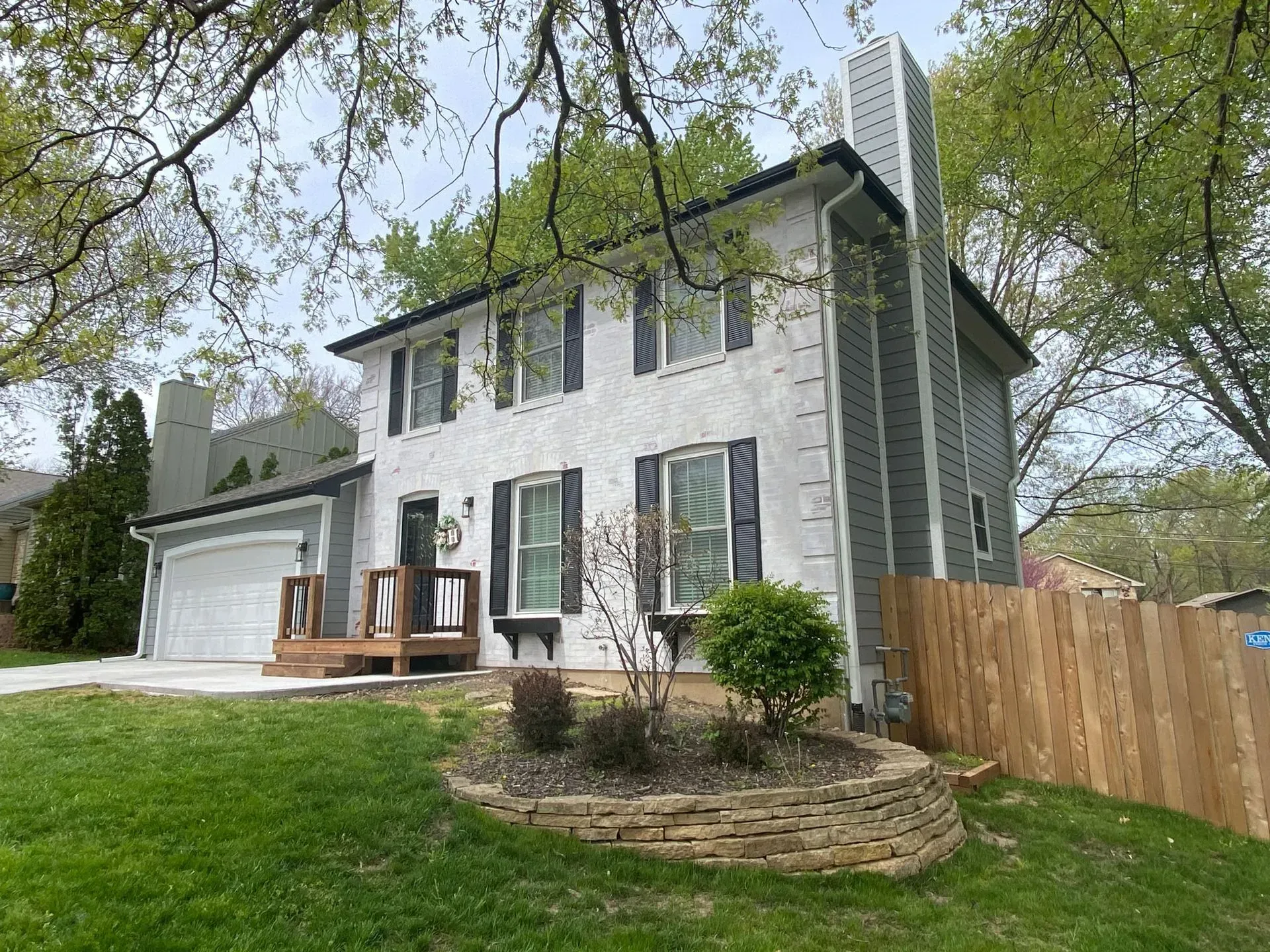 A two-story white house with dark shutters and a grey chimney, featuring a stone retaining wall in the front yard.
