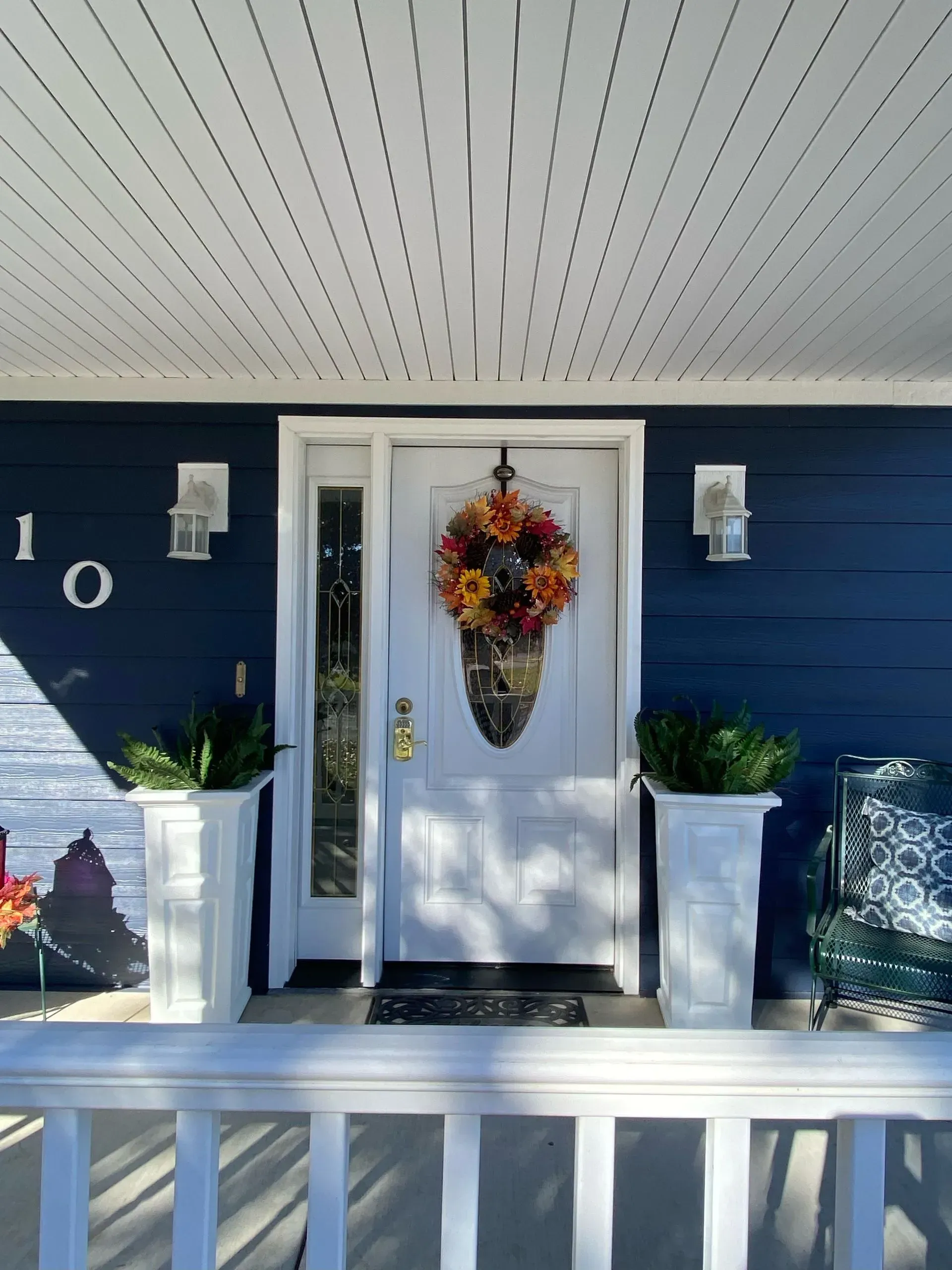 A white front door with a fall wreath, flanked by white planters and dark blue siding, viewed from a porch.