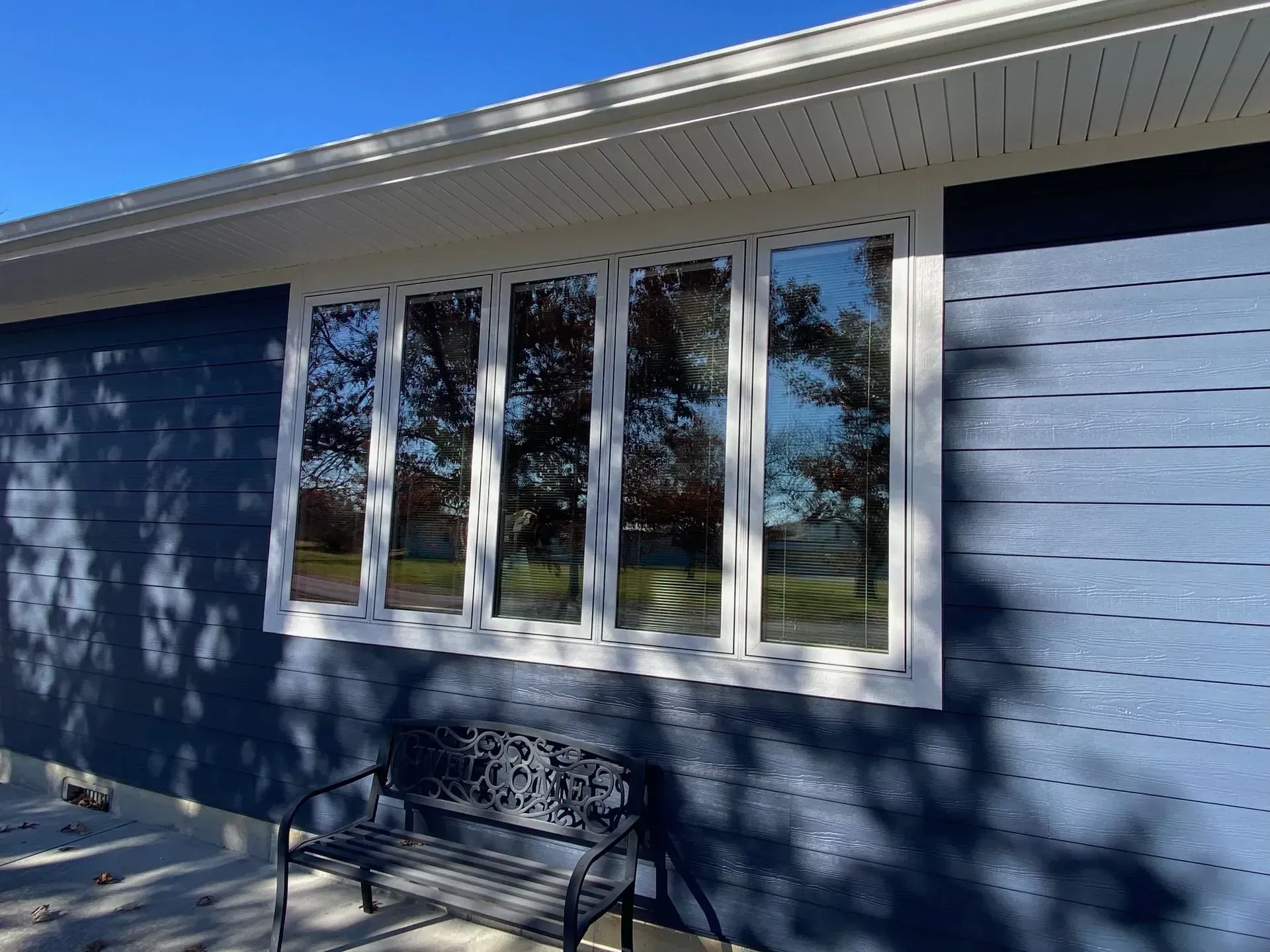 A large four-pane window with a white frame on a dark blue horizontal-sided house wall, with a metal bench underneath.