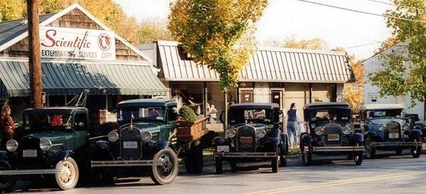 A row of vintage cars parked along a street in front of a rustic storefront on a sunny autumn day.