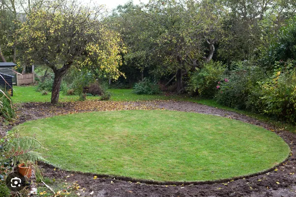 Oval green lawn in a garden, bordered by shrubs and trees on a cloudy day