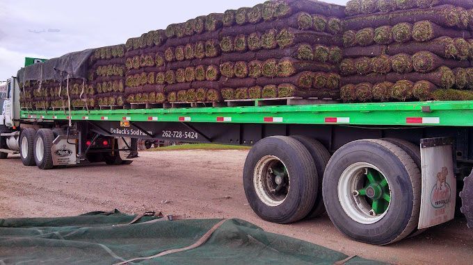Flatbed truck loaded with stacked brown cargo rolls along a gravel road.