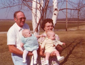 Two adults holding babies outdoors in a sunny field near bare trees and farmland