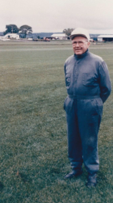 Person in a blue jacket and cap standing on a grassy field with buildings in the background.