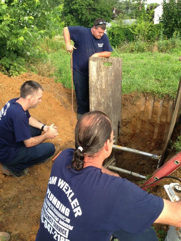 A man wearing a wexler plumbing shirt is kneeling in the dirt.w