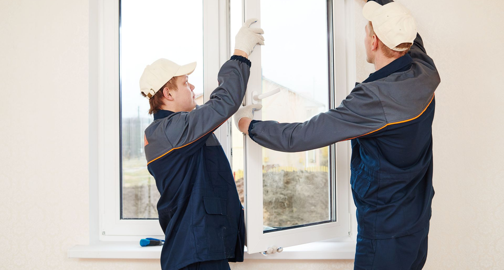 Two workers installing a window frame indoors, wearing white gloves and navy uniforms.
