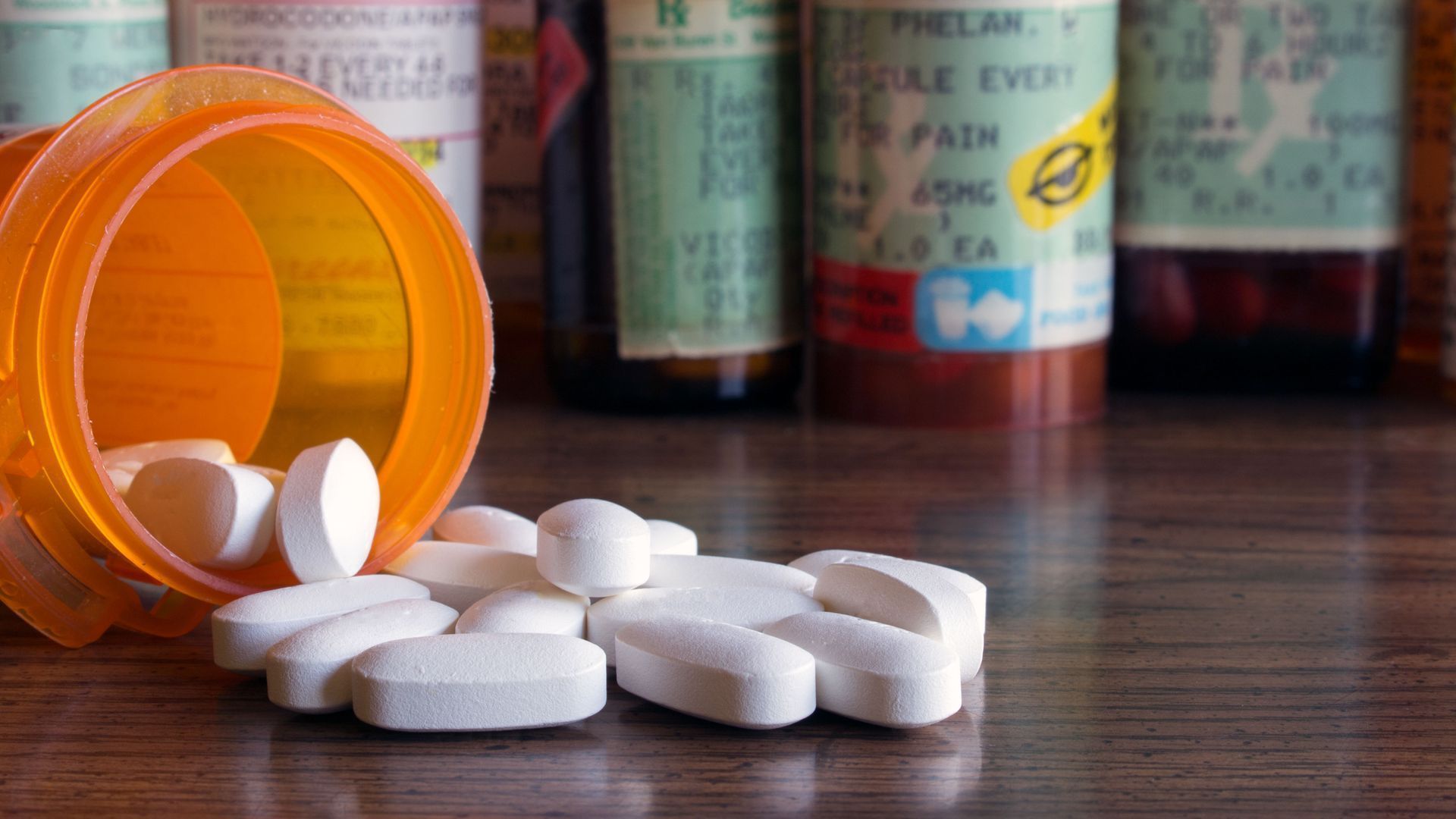 Orange pill bottle tipped over with white tablets spilled on a wooden surface, blurred medicine bottles in back