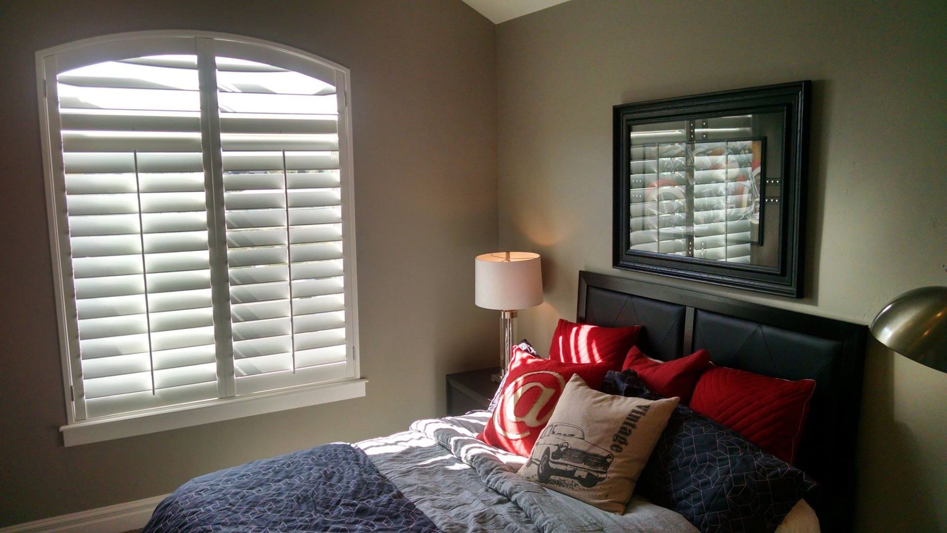 Bedroom with arched window and white shutters, bed with red and blue pillows, mirror on the wall.