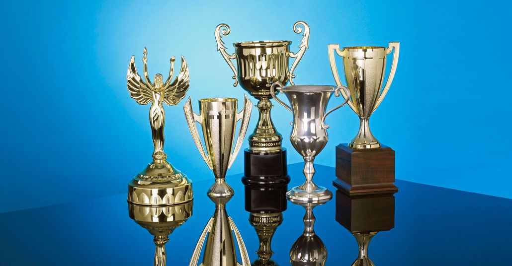 Six gold and silver trophies on a reflective surface, against a blue gradient background.