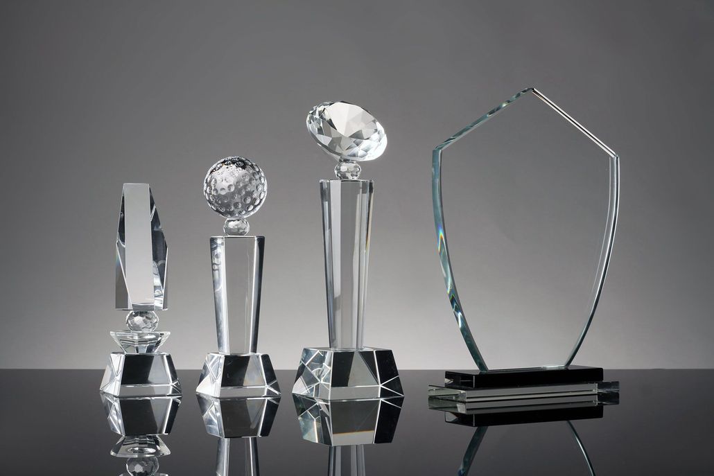 Glass trophies on a reflective surface, ranging in shape and size, set against a gray backdrop.