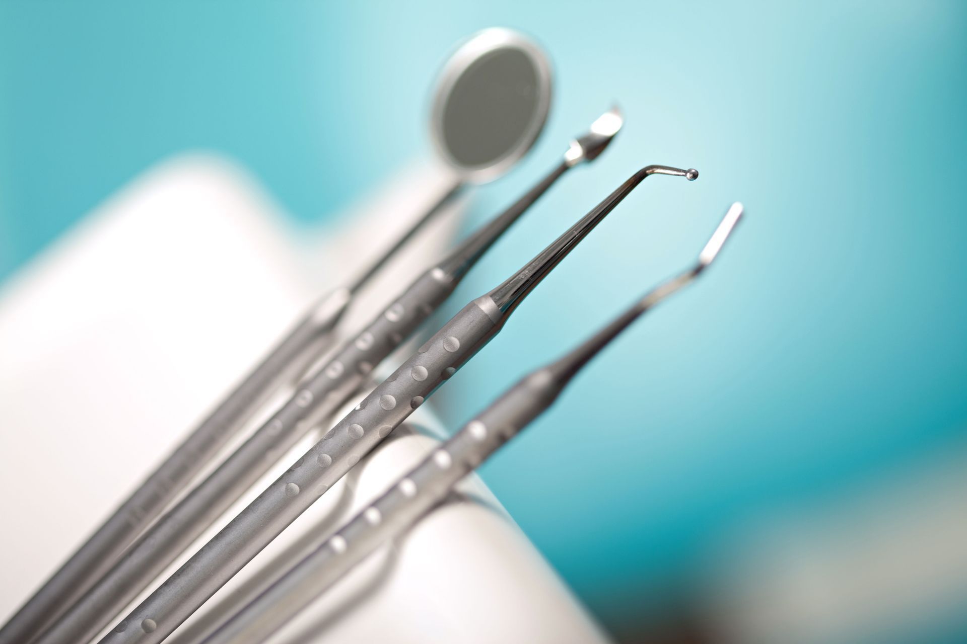 Dental tools on a white tray: mirror, probe, explorer, and scaler. Blue background.