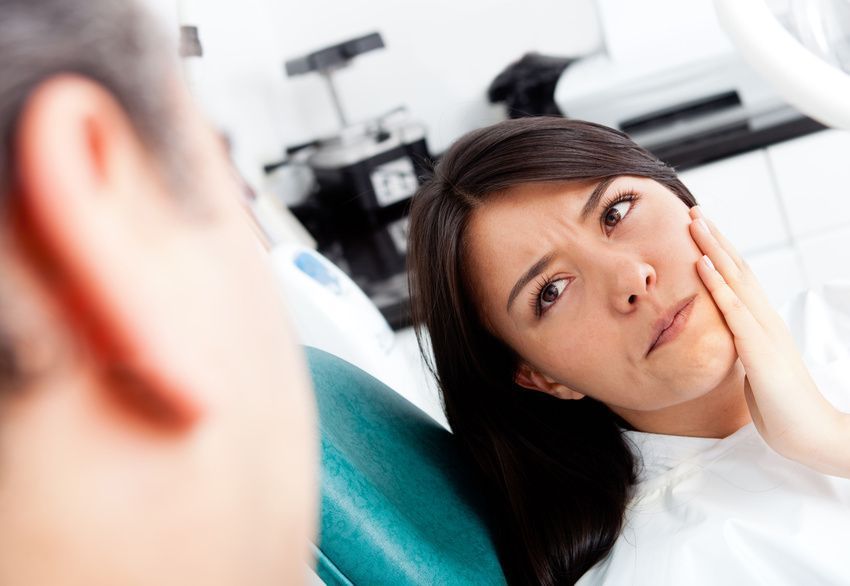 Woman in dentist chair, holding cheek in pain, looking concerned. Medical setting.
