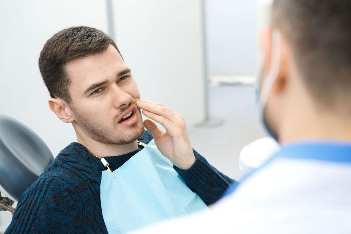 Man at dentist, holding jaw, appearing to be in pain. Blue bib, doctor in background.