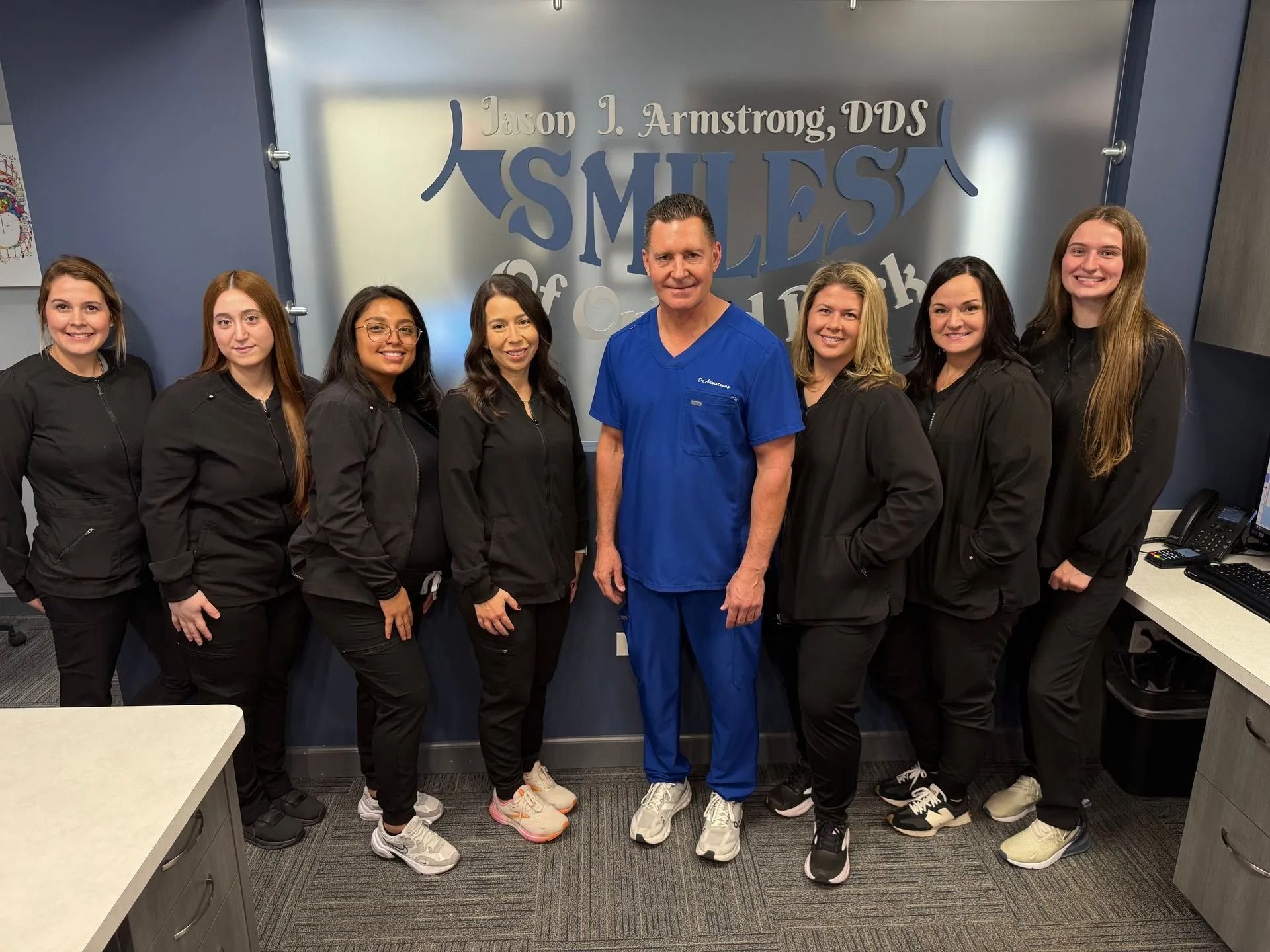 Staff poses in dental office; man in blue scrubs stands with women in black. Logo on glass.