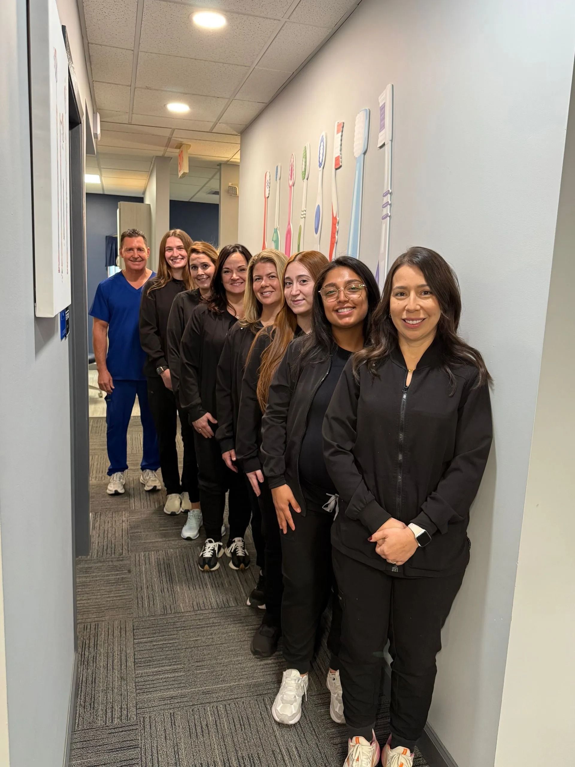 A dental team in black scrubs lines up in a hallway, smiling, with one person in blue scrubs.