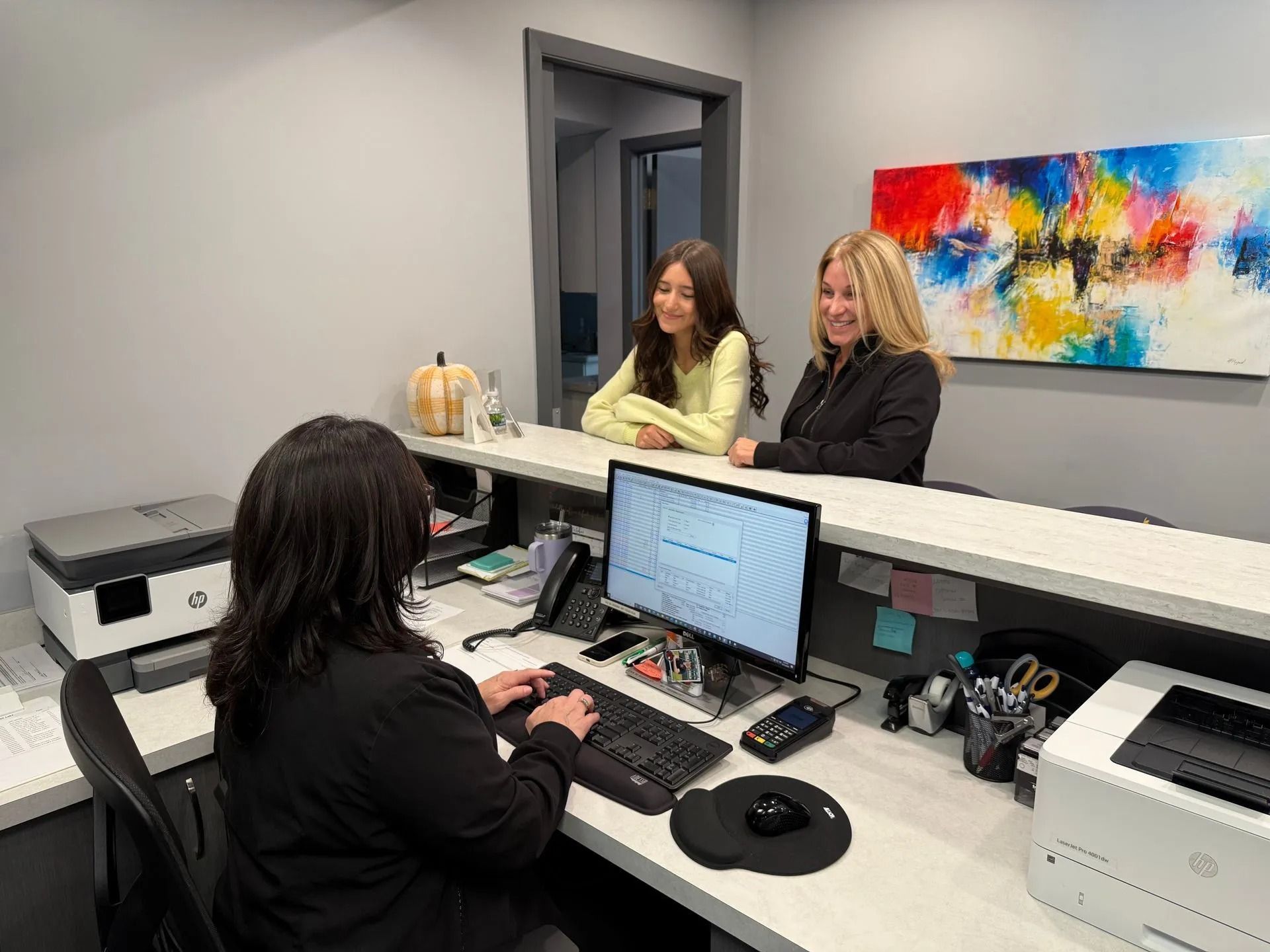 Receptionist assisting two people at a desk in an office. Colorful artwork hangs on the wall.