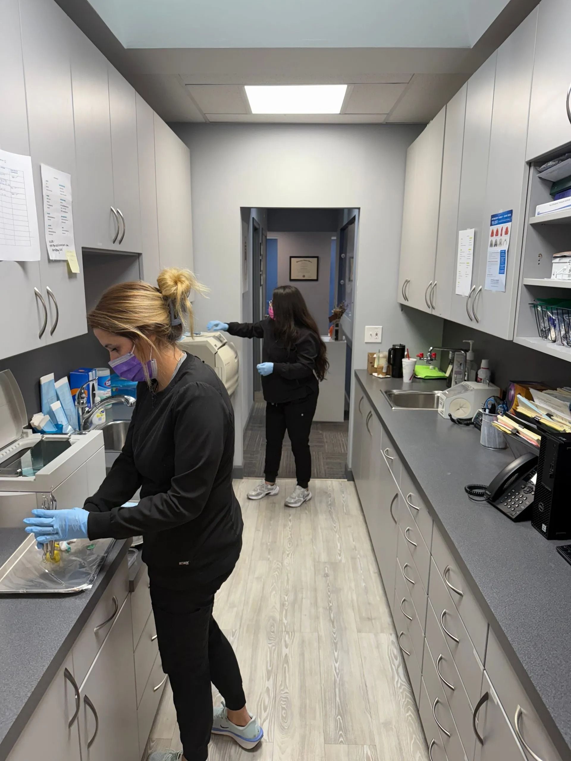 Two dental assistants in a narrow office, wearing masks and gloves, preparing equipment.