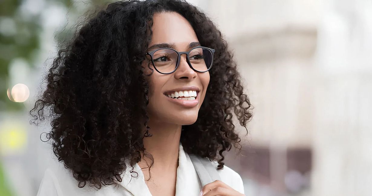 Woman with curly hair and glasses smiles outdoors.