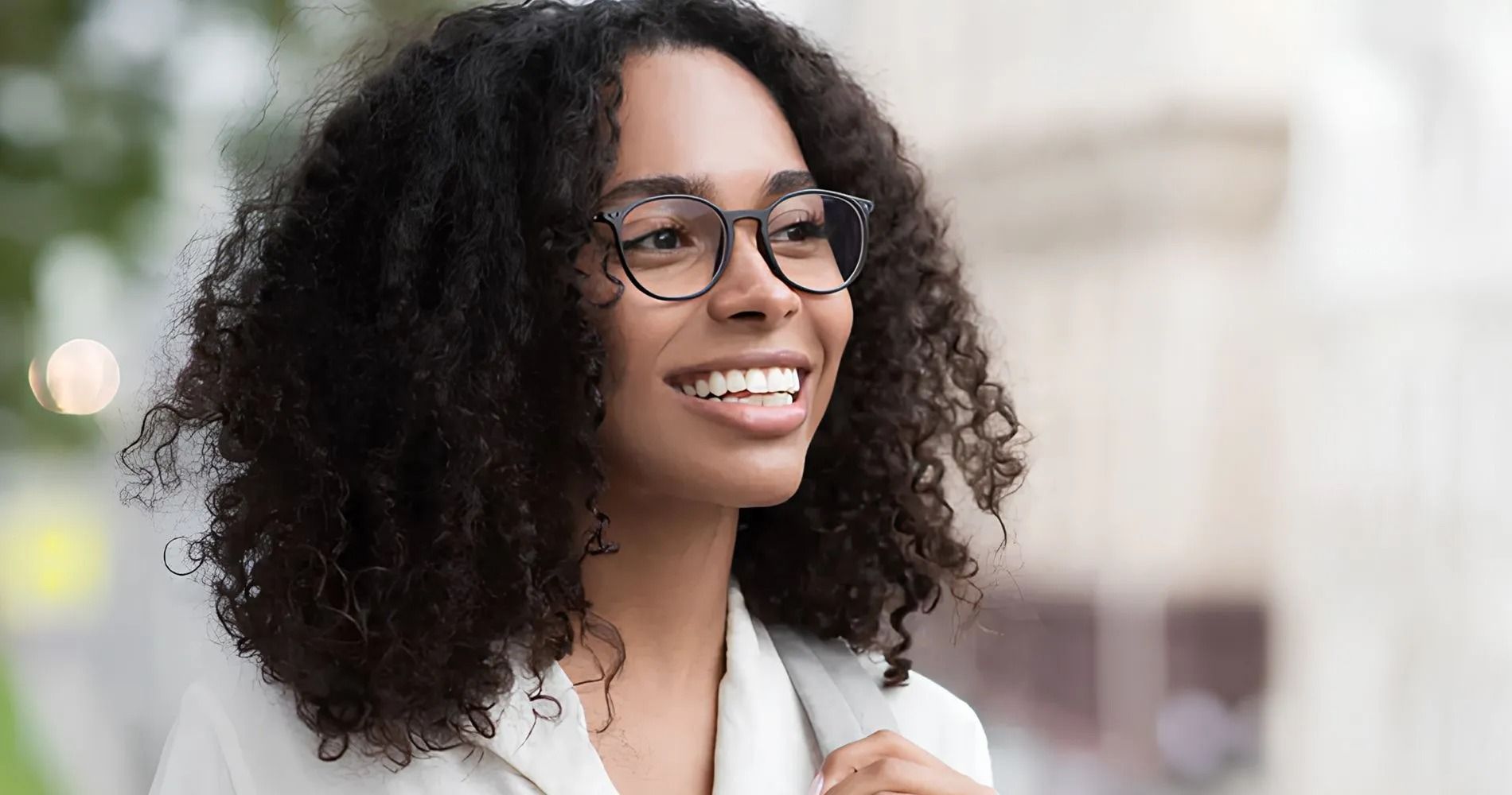 Woman with curly hair and glasses smiles outdoors.