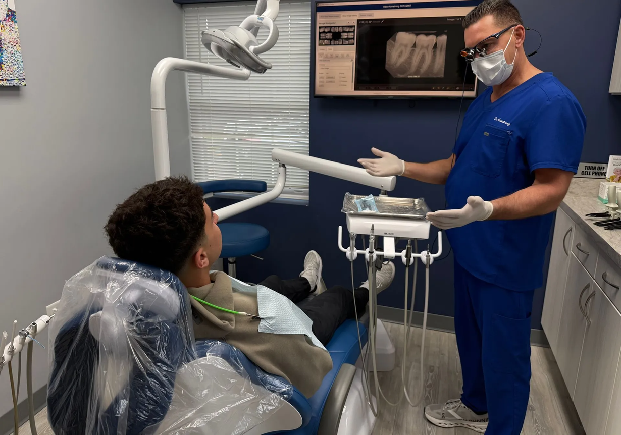 Dentist in blue scrubs points to dental tools. Patient in chair, dental office.
