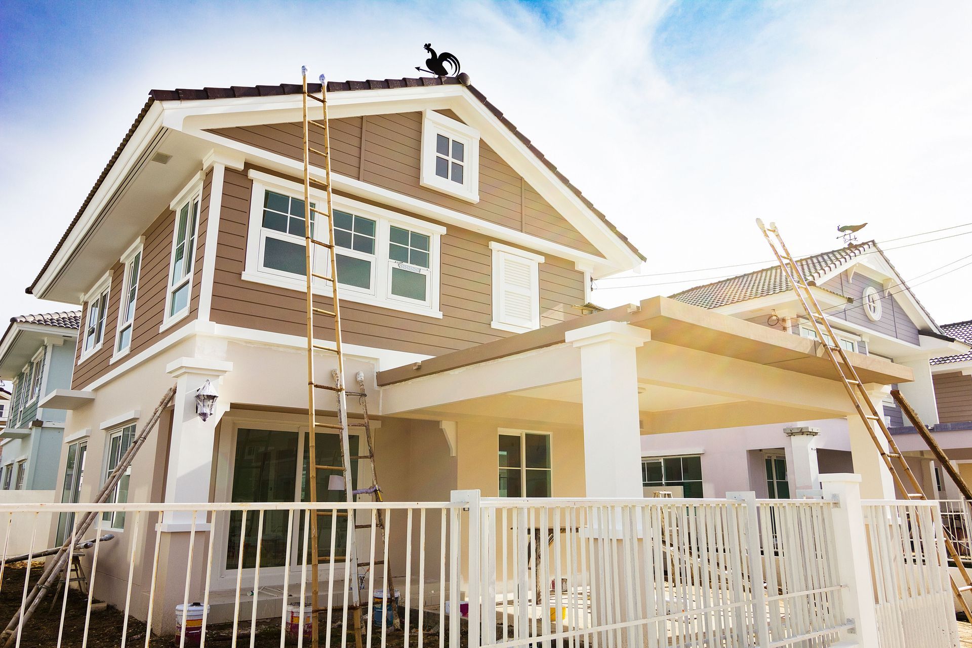 Two-story house under construction, brown siding, white trim, worker on the roof, ladder, sunny day.