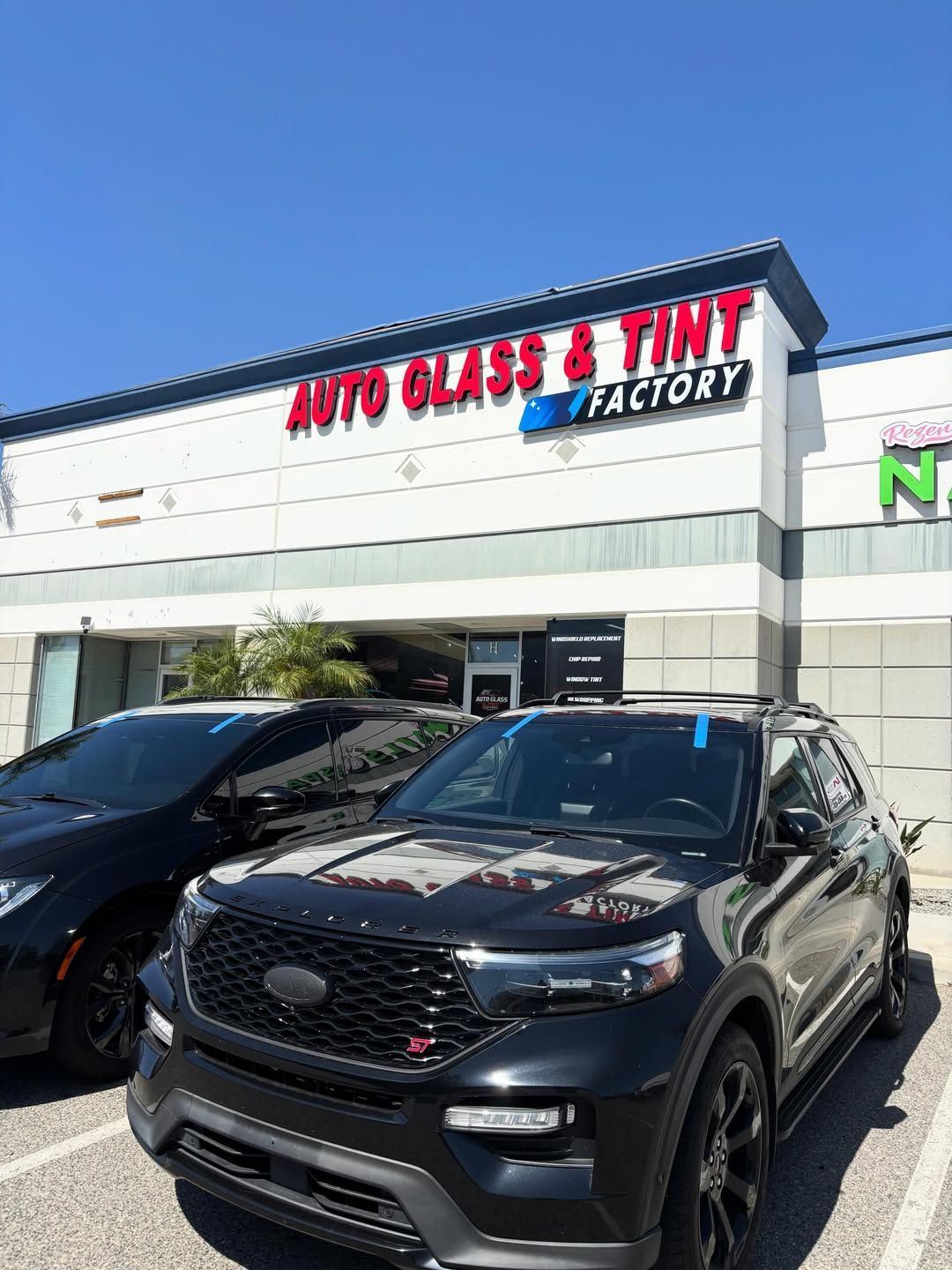 Two black SUVs parked outside an Auto Glass & Tint Factory storefront under a clear blue sky