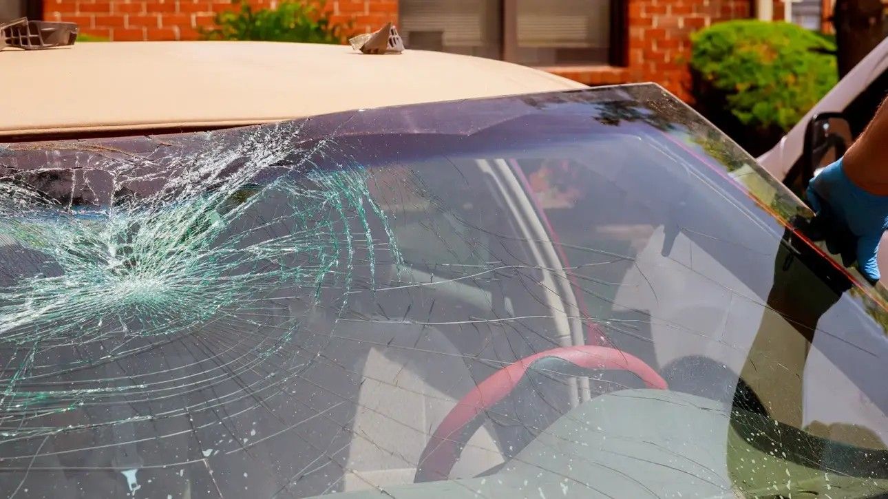 Cracked windshield on a car parked outside a brick building, with shattered glass across the front.