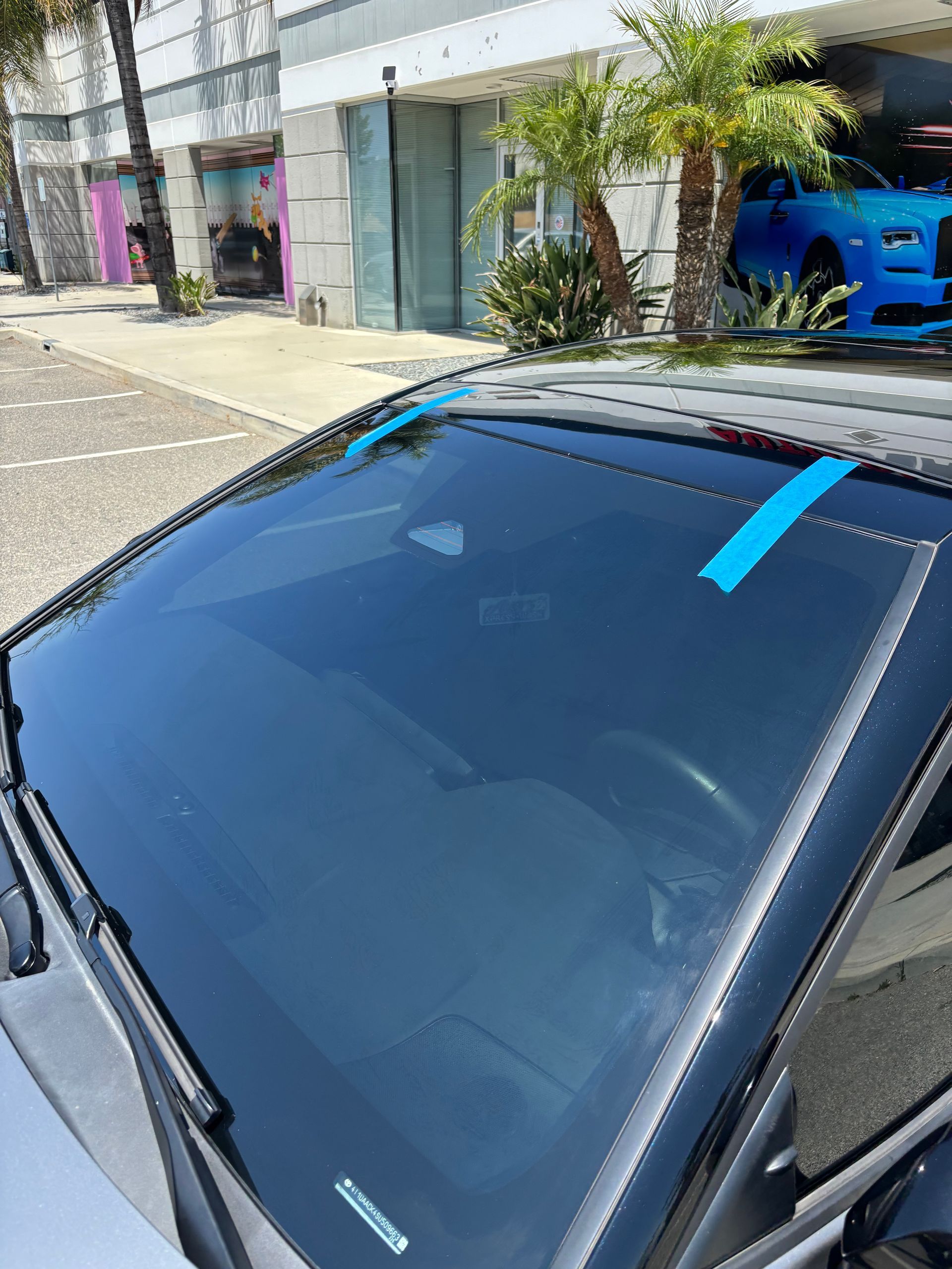 Blue car parked outside storefronts, with a bright blue tape strip on the windshield.
