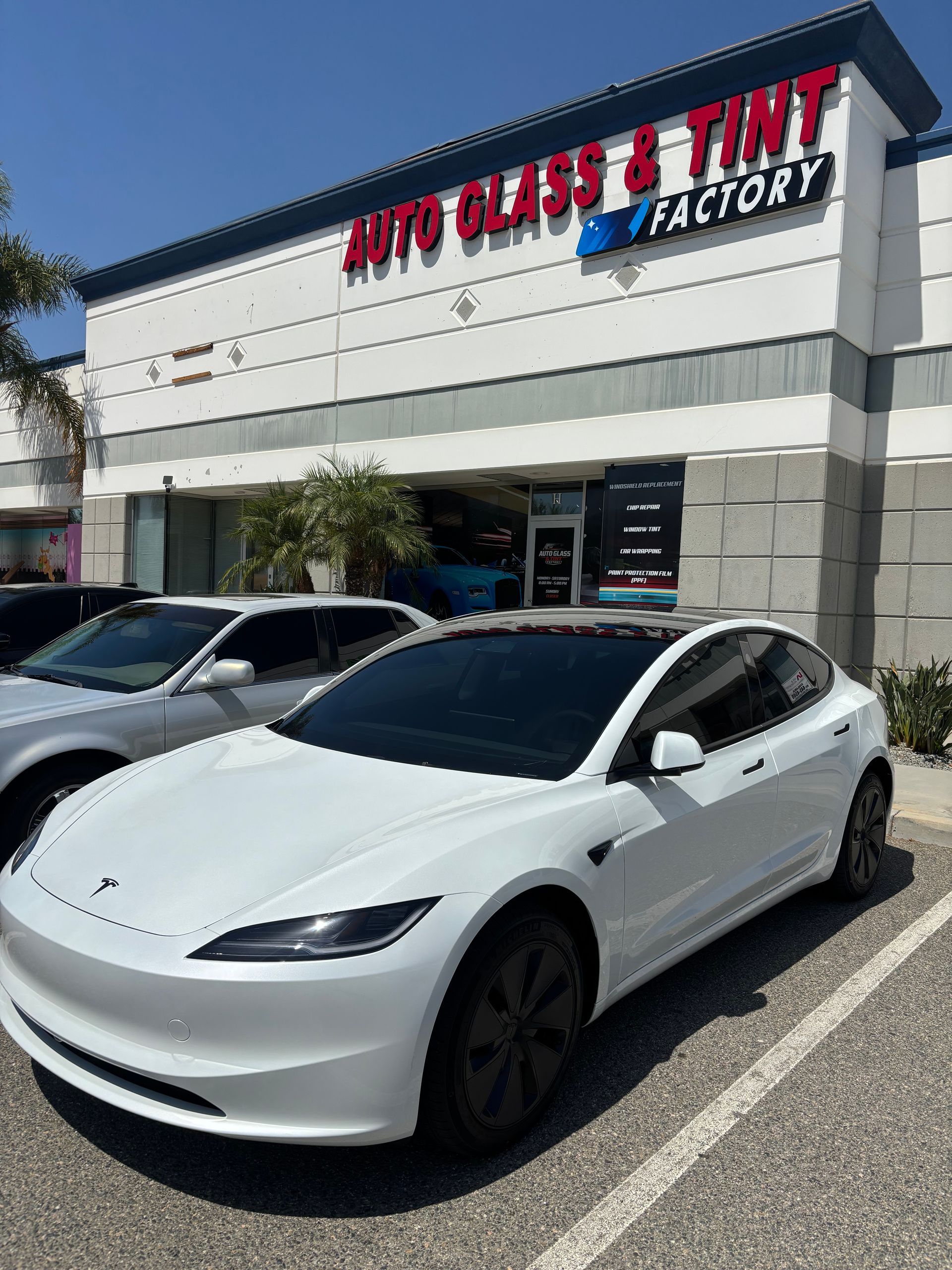 White Tesla sedan parked outside an auto window tint shop with red and blue signage.