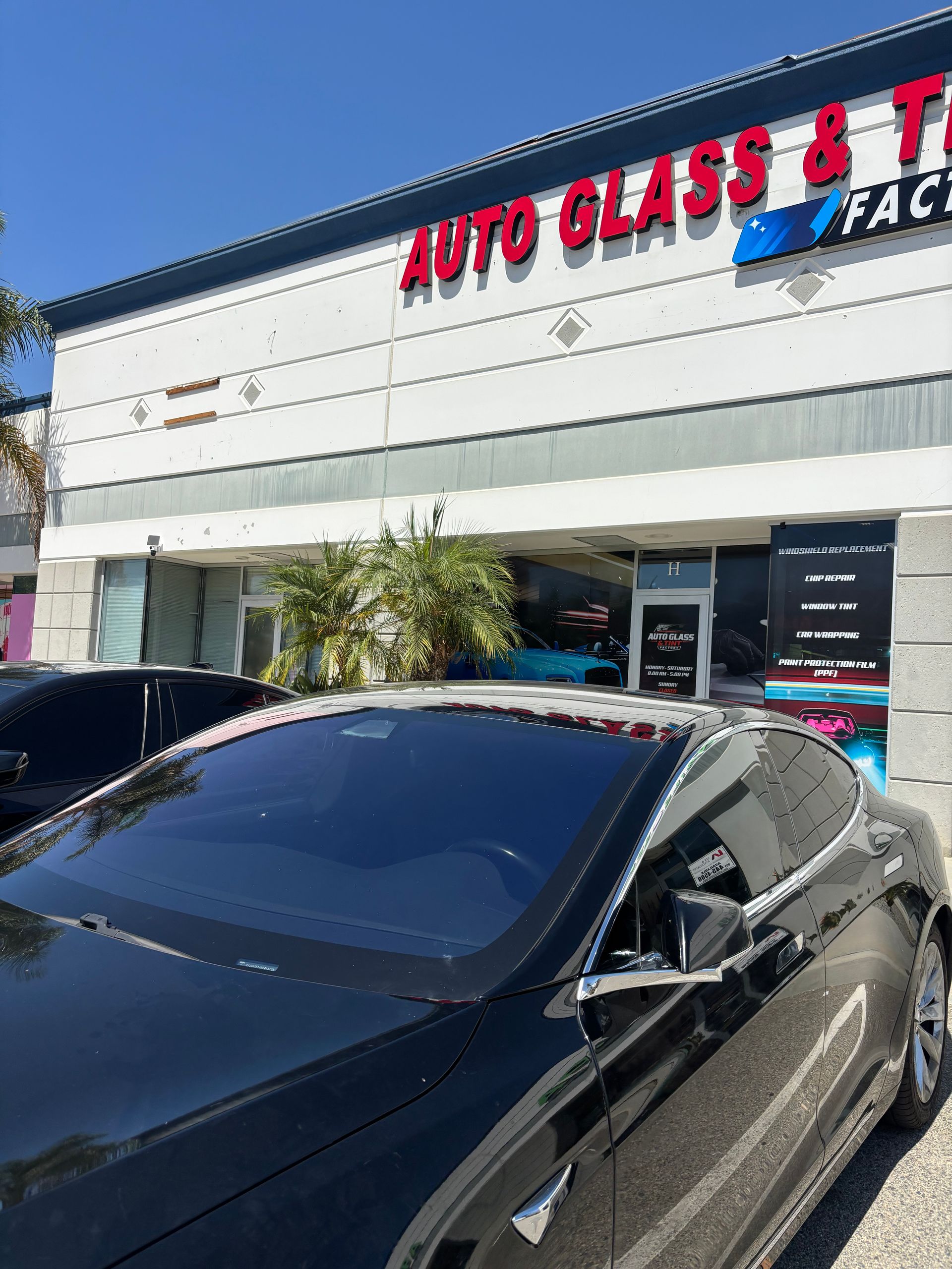 Black car parked outside an auto glass and tint shop under bright sunlight
