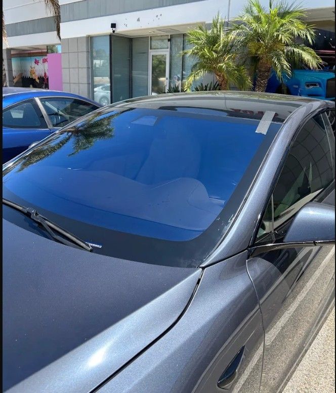 Silver car parked outside a storefront with a large blue-tinted windshield under bright sunlight