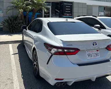 White Toyota sedan parked outside a building with palm trees and another car nearby.