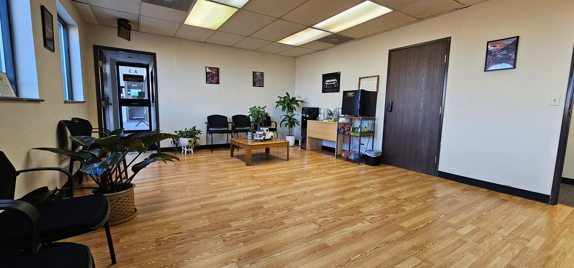 Empty waiting room with wood floor, chairs, plants, posters, and a closed door