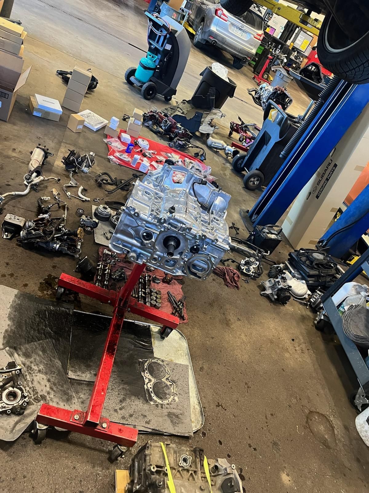 Car engine block on a red stand in a cluttered auto repair shop floor, with parts scattered around.