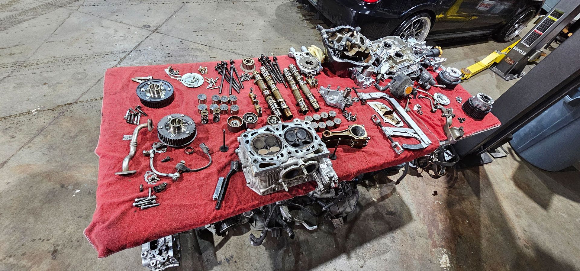 Disassembled car engine parts spread on a red mat in a garage repair area.