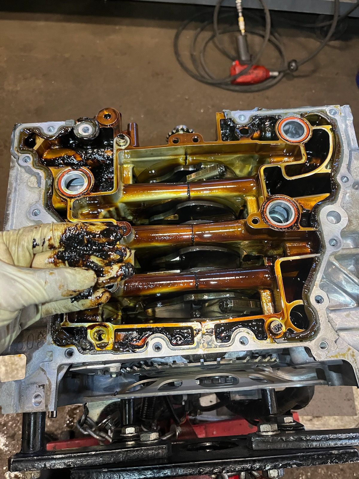 Gloved hand holding an oily engine valve cover with gasket residue and exposed engine parts underneath