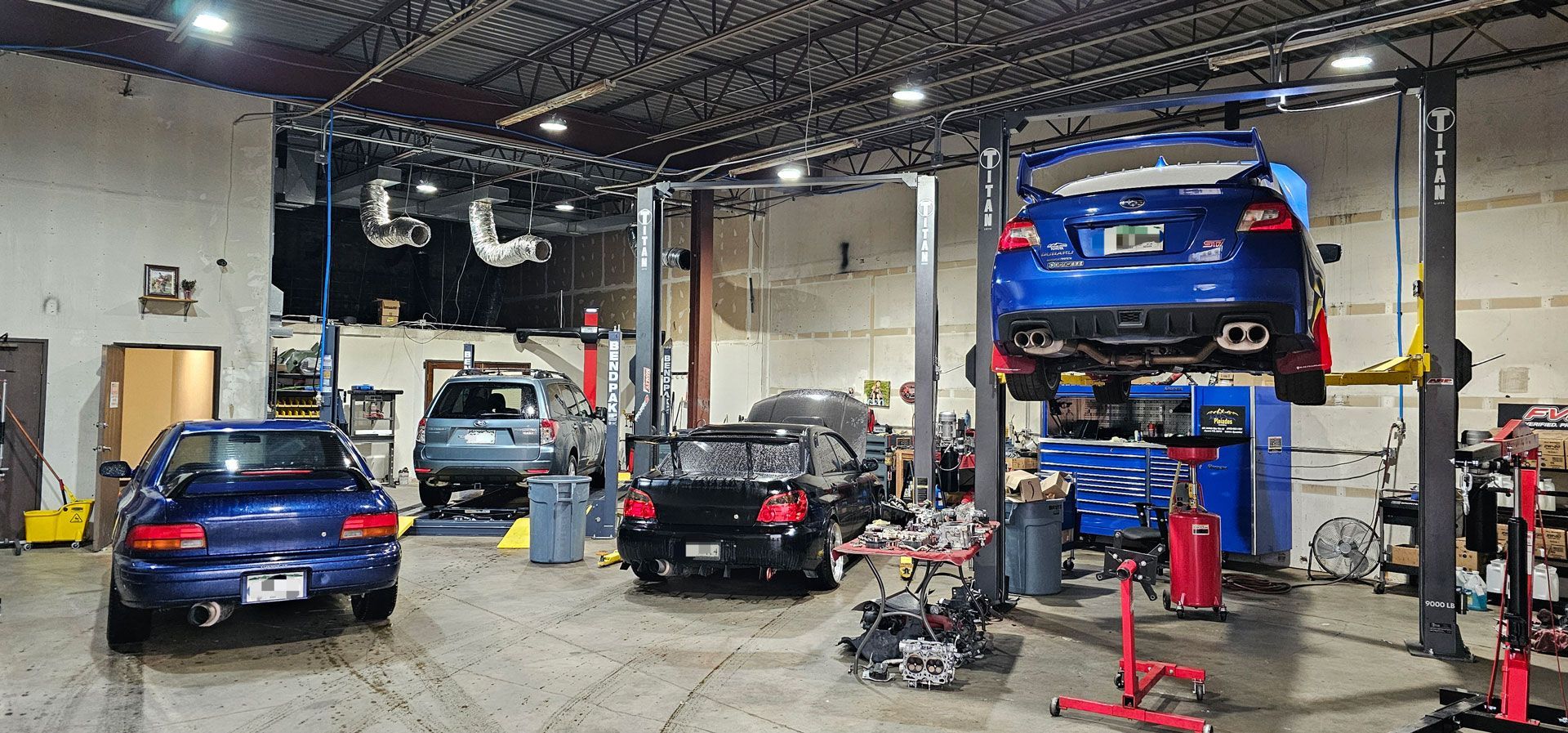 Auto repair shop with several cars on lifts, including a blue car raised on a rack and another on the floor.