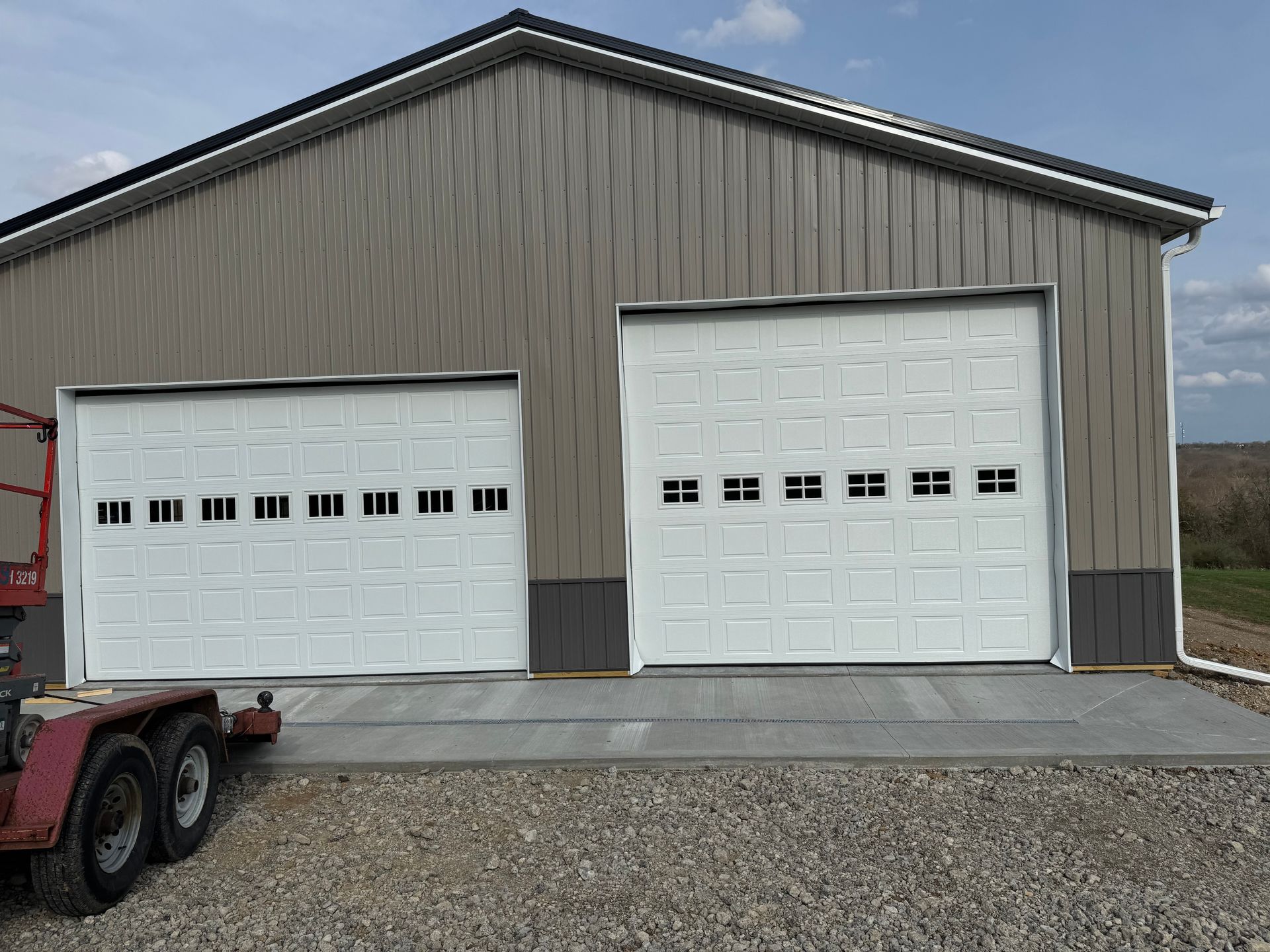 A large garage with two white garage doors and a red trailer parked in front of it.