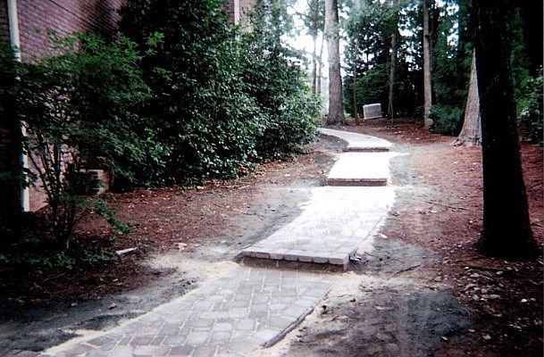 A brick walkway is surrounded by trees and dirt.