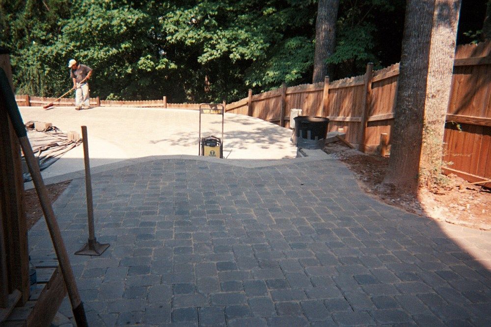 A man is laying bricks on a patio next to a wooden fence.