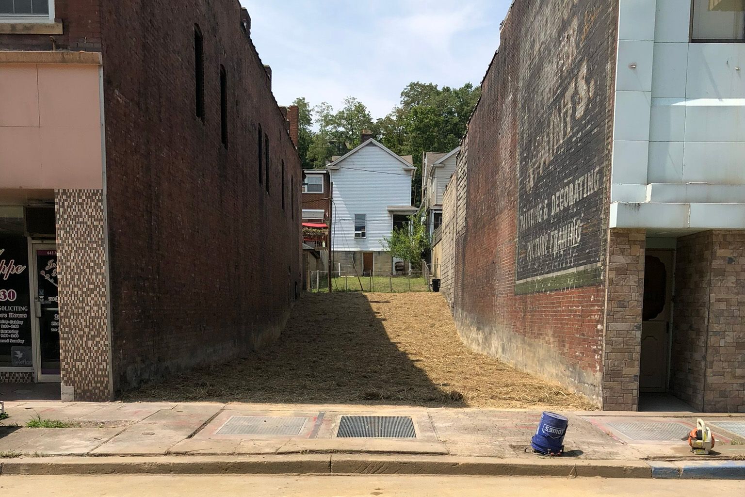Narrow vacant lot between brick buildings, revealing a house in the distance.