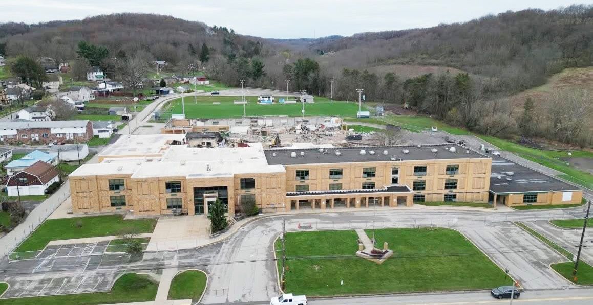 Aerial view of a light-brown brick building, possibly a school or hotel, with a partial roof replacement, in a hilly, rural setting.