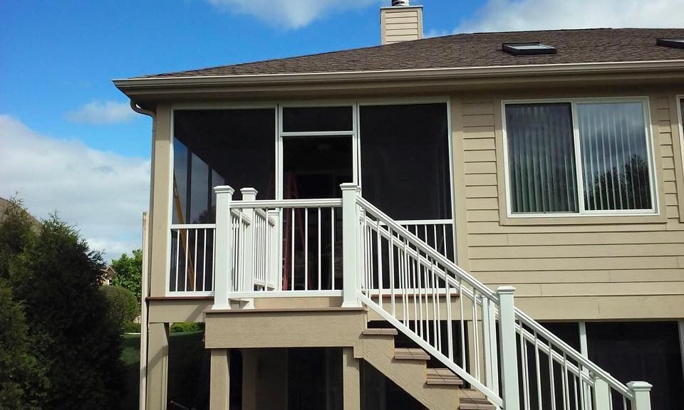 Screened-in porch attached to a beige house, with white railings and stairs, blue sky in the background.