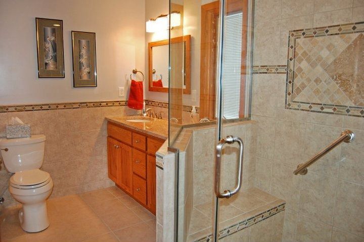 Bathroom with glass shower, toilet, vanity, and grab bar. Light beige tiles and wood cabinets.