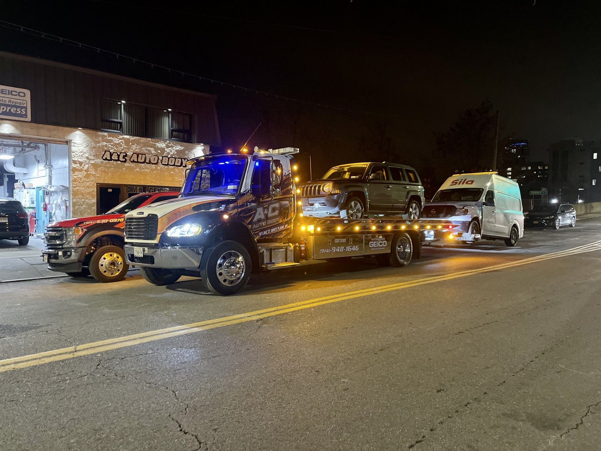 A tow truck is towing a car down a street at night.