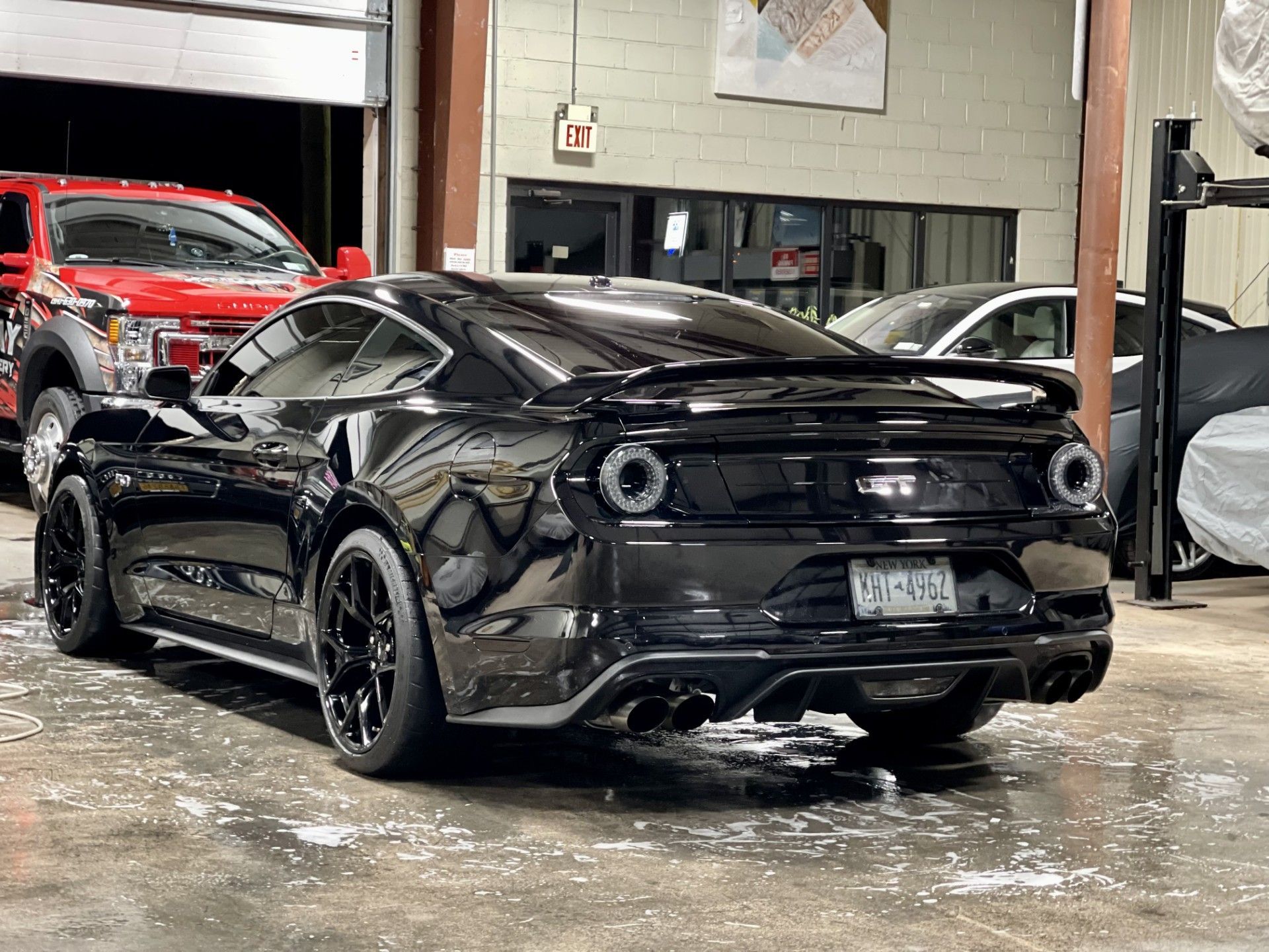 A black mustang is parked in a garage next to a red truck.