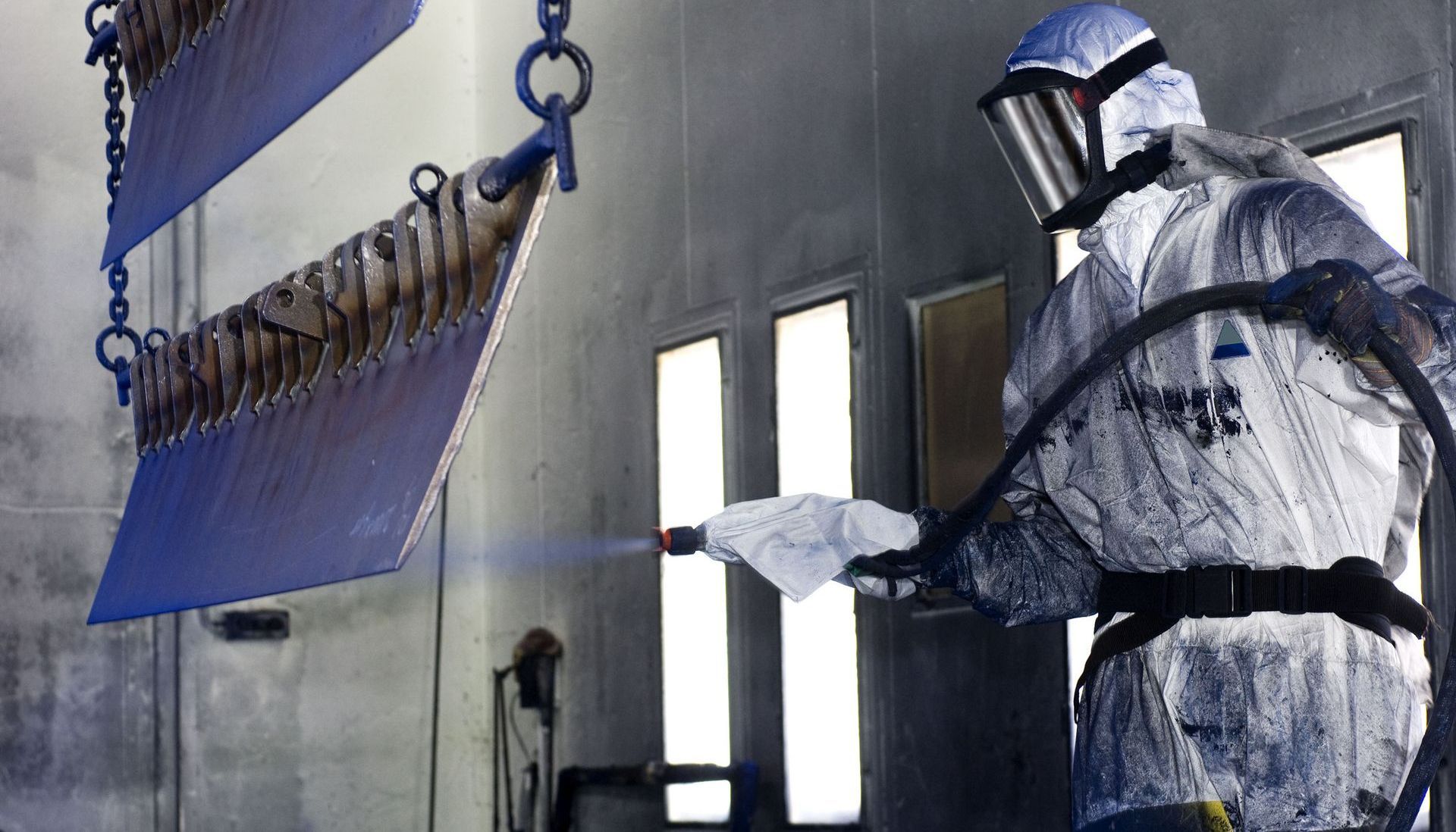A person in protective gear spray-painting metal objects inside a paint booth.
