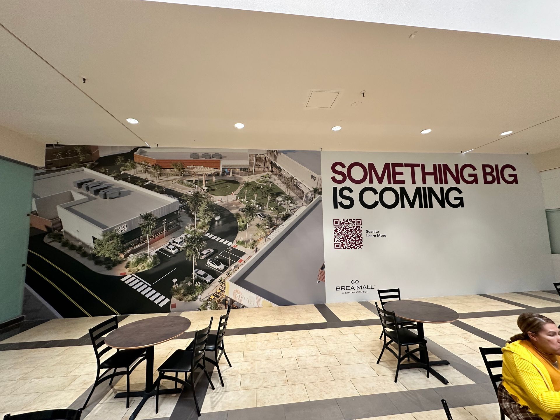 A woman sits at a table in front of a wall that says something big is coming